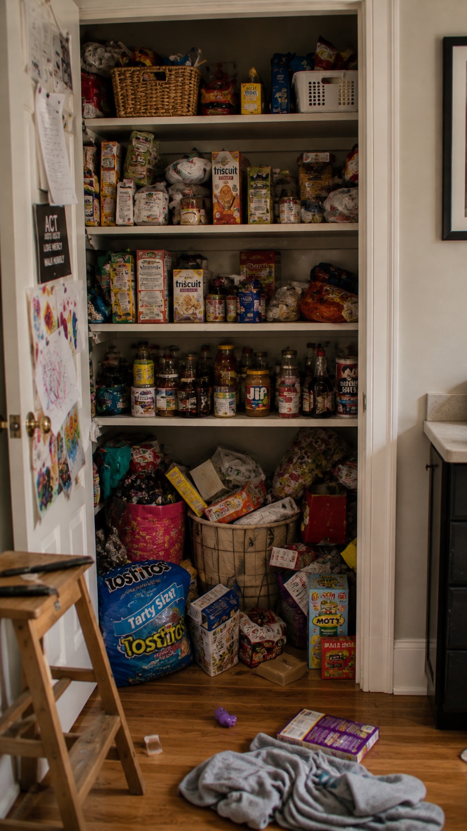 A cluttered pantry before organizing — overflowing shelves, mismatched boxes, and a stool tucked beside the chaos