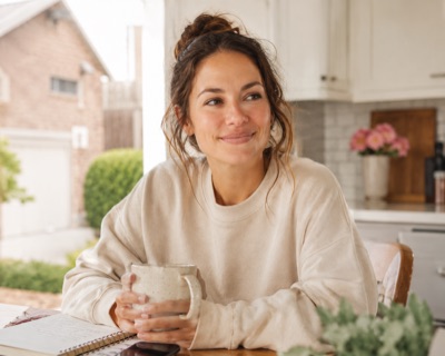 Rachel M., a brunette in her thirties in a cream sweater holding a coffee mug at her kitchen table